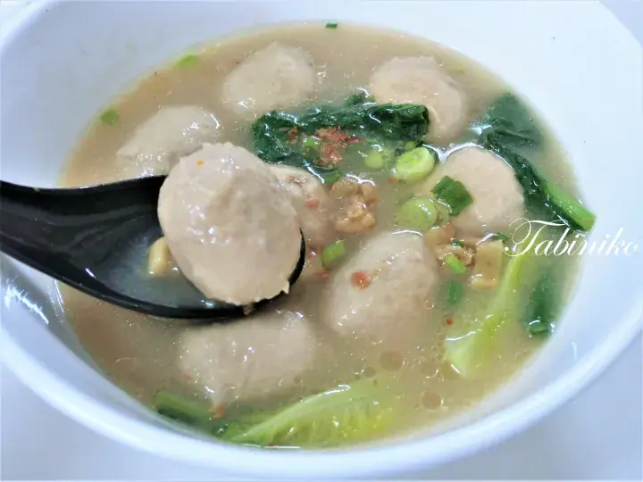 A close-up of a black spoon lifting a fish ball from a bowl of clear soup with greens and minced pork.