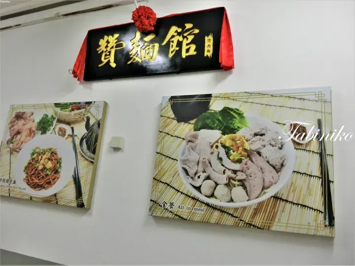 A black sign with gold Chinese characters reading Zan Mian Guan hangs above wall-mounted photos of noodle dishes at a restaurant.