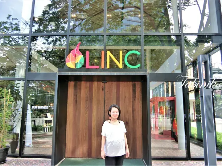 A woman stands smiling in front of the entrance to LINC, a modern glass-fronted building with a colorful logo.