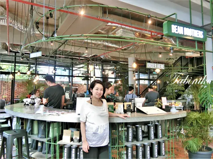 A woman stands at the counter inside Bean Brothers cafe, featuring industrial decor with green metal frames and hanging lights.