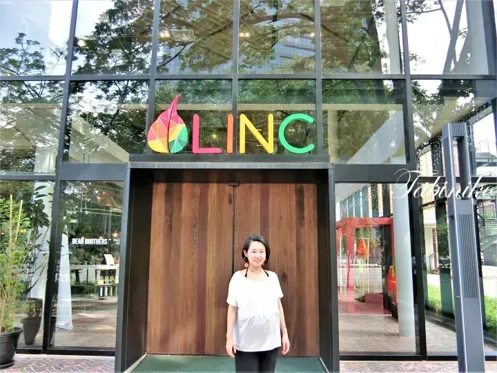 カラフルなロゴが目立つガラス張りのモダンな建物「LINC」の前に立つ女性。