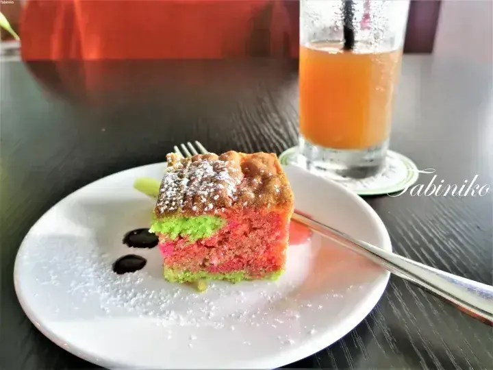 緑とピンクの層が特徴的なケーキが粉糖をまぶして白い皿に盛り付けられ、隣にはアイスジュースが添えられています。