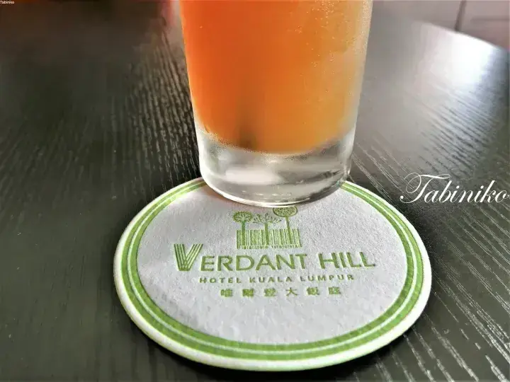 ダークウッドのテーブルに置かれた、Verdant Hill Hotel Kuala Lumpurのコースターの上のオレンジジュース。