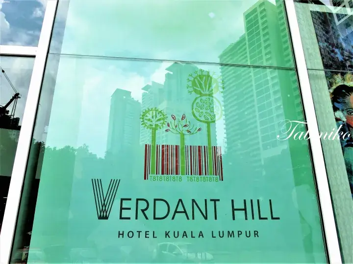 Glass facade of Verdant Hill Hotel Kuala Lumpur featuring a green logo with trees and a barcode design.
