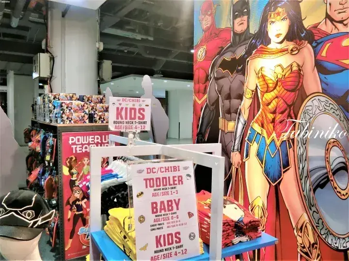 ワンダーウーマンやバットマンなどのDCコミックグッズと子供服のサイズ表示が並ぶ商品陳列。