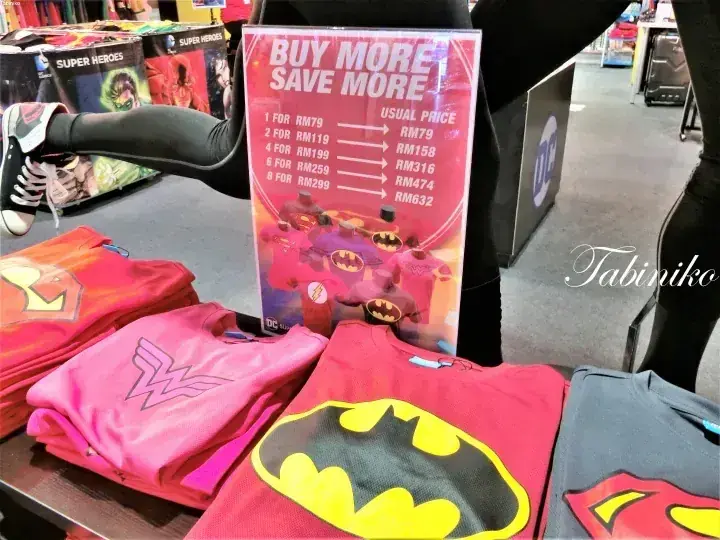 「BUY MORE SAVE MORE」の価格表と折り畳まれたTシャツが並ぶDCコミックスのスーパーヒーローグッズの店頭陳列。