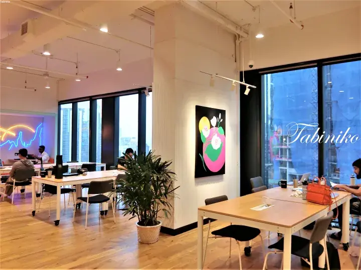 Modern co-working space with wooden floors, large windows, and a colorful floral artwork on the wall.