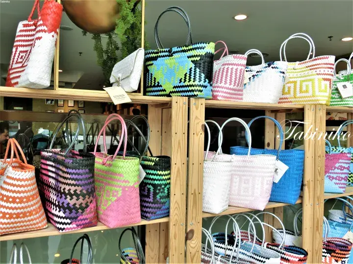 Colorful woven tote bags with geometric patterns displayed on wooden shelves at a Tabiniko store.