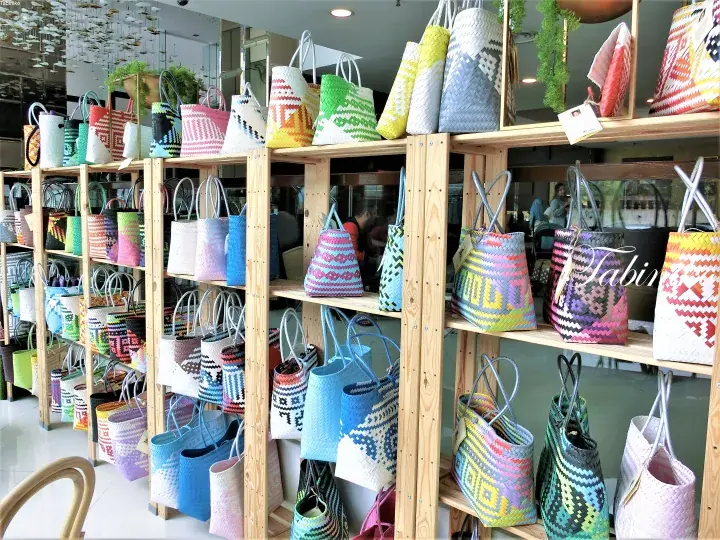 Colorful woven tote bags displayed on wooden shelves in a bright retail store.