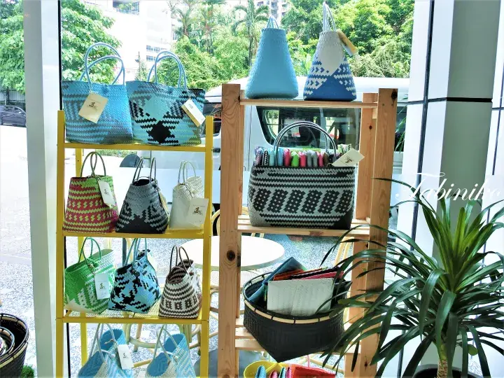 Colorful woven bags displayed on yellow and wooden shelves in a bright shop window with a plant.