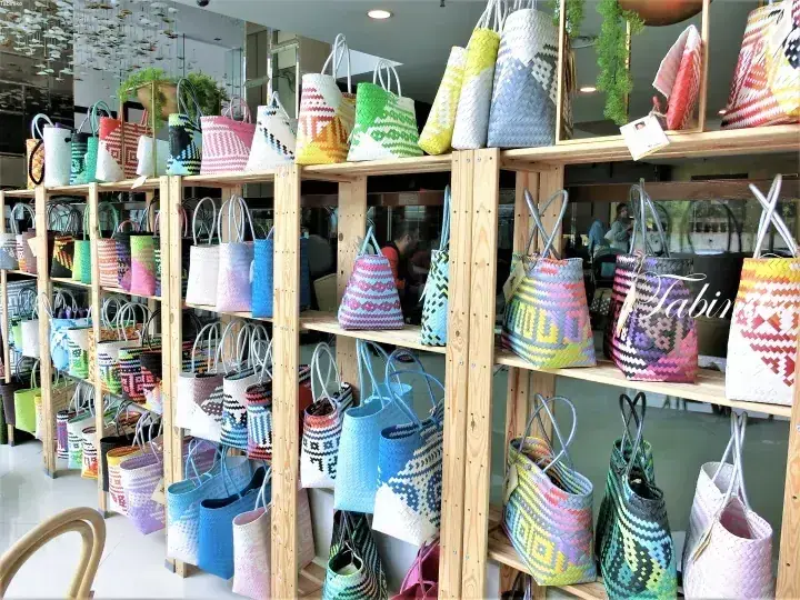 明るい店内の木製棚に並ぶカラフルな織りトートバッグ。