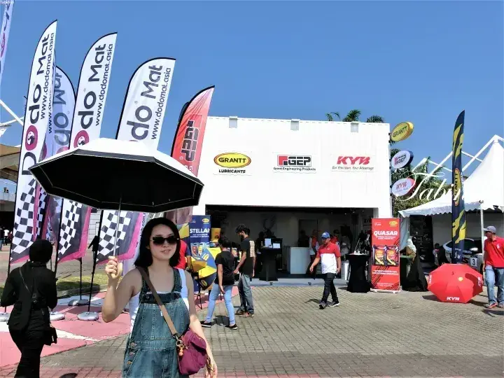 GRANTTやKYBのバナーが並ぶ屋外自動車展示会で黒い傘をさす女性。