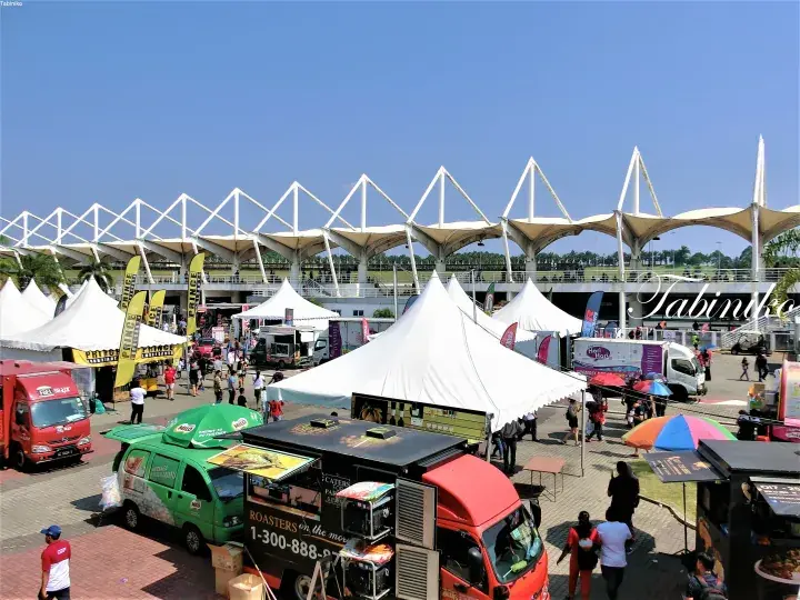 Food trucks and white tents at an outdoor event with a modern white tensile structure roof in the background.