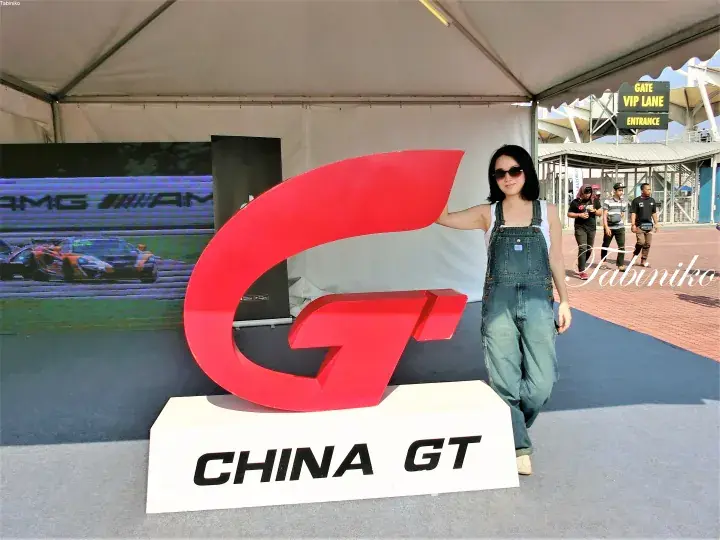 A woman poses beside a large red China GT logo at a motorsport event entrance.