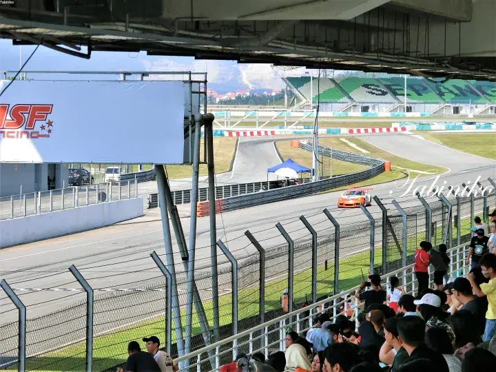Orange race car speeding on a track at Sepang International Circuit with spectators watching from the stands.