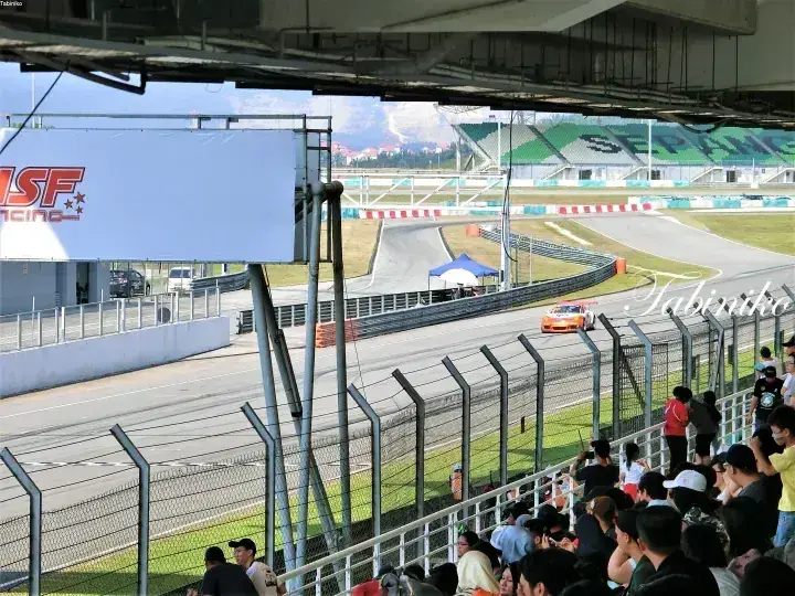 観客が見守る中、セパン・インターナショナル・サーキットを疾走するオレンジのレーシングカー。