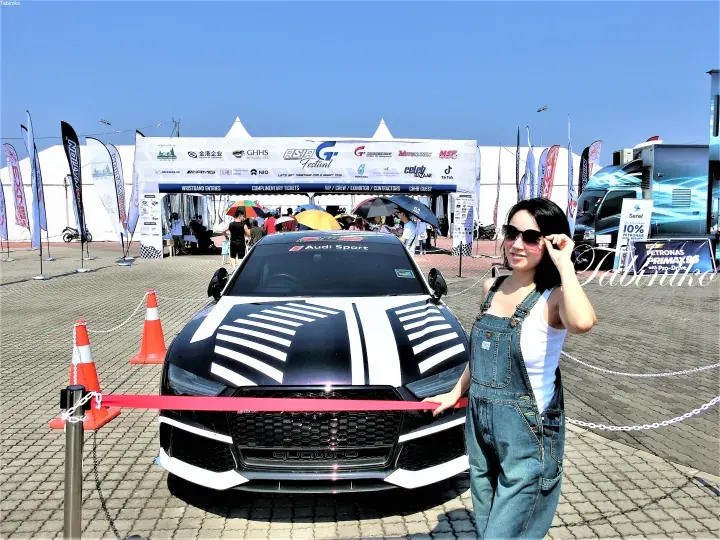 Woman posing beside a black Audi Sport car with white stripes at an outdoor automotive event.