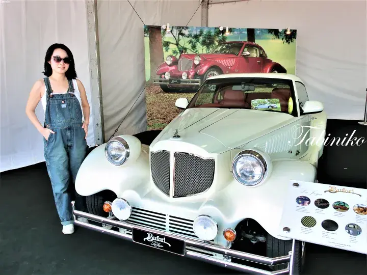 A woman in denim overalls stands beside a white vintage Bitter car at an exhibition.