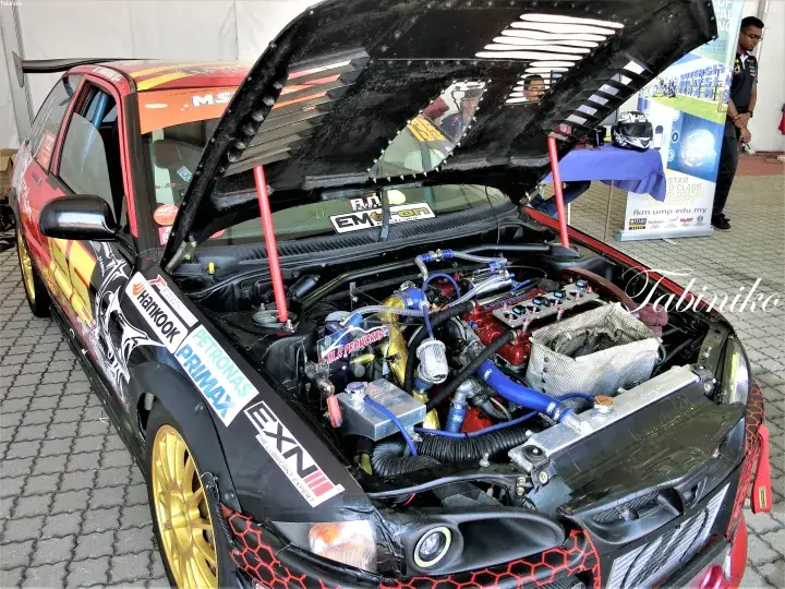 Modified car with open hood showing engine bay and sponsor decals like Hankook and Petronas at a Malaysian car show.