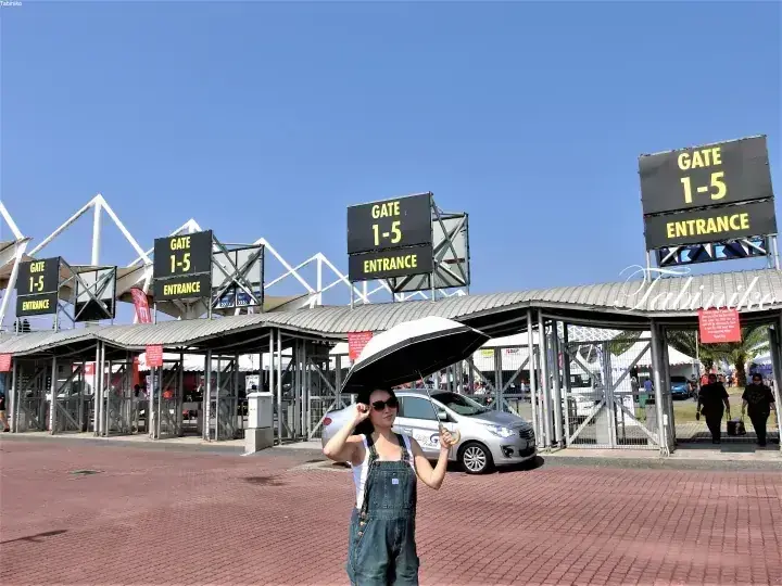 晴れた空の下、大きな屋外施設のGate 1-5入口前に傘をさした女性が立っている。