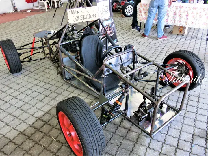 Custom-built go-kart chassis with red wheels and a black seat displayed at an indoor exhibition.