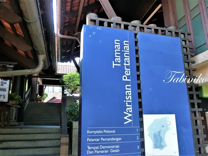 Blue signboard for Taman Warisan Pertanian listing visitor complex, viewing platform, and demonstration areas.