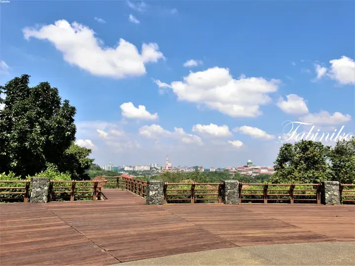 Wooden viewing deck with stone pillars overlooking a cityscape under a blue sky with white clouds.
