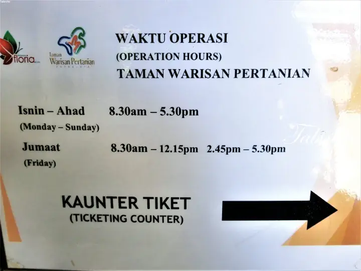Sign showing operation hours for Taman Warisan Pertanian, with ticketing counter direction.