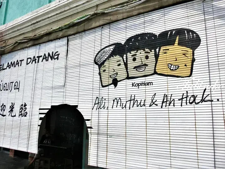 「Selamat Datang」の文字と「Ali, Muthu & Ah Hock」の顔の壁画が描かれた店舗のシャッター。