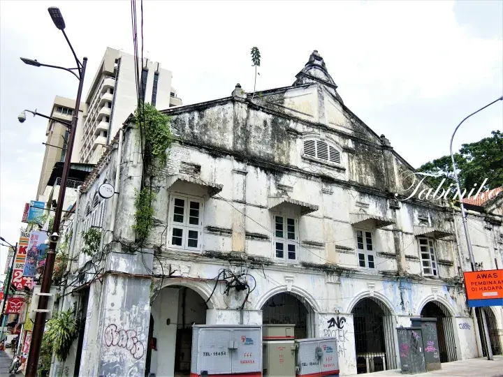 Weathered colonial shophouse facade with arched ground floor and graffiti, set against modern high-rise buildings in an urban Malaysian street.
