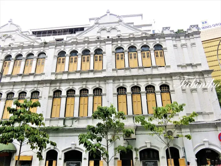 White colonial-style shophouse with yellow wooden shutters and arched windows, featuring trees in front.