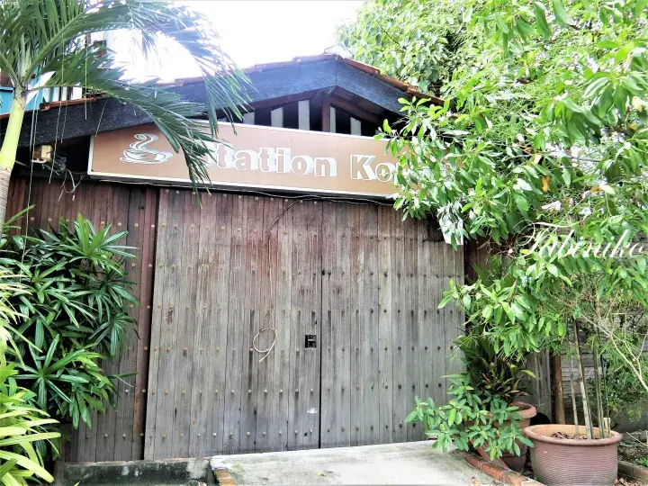 Wooden gate of Station Kopi with a sign featuring a coffee cup logo, surrounded by lush greenery.