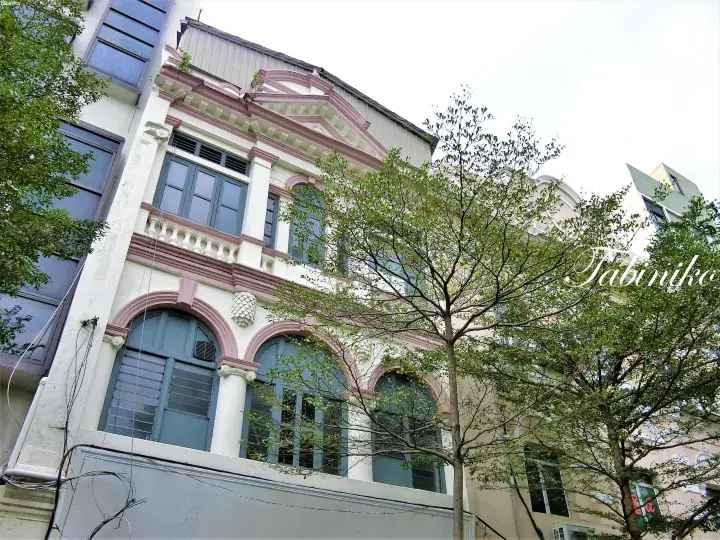 A low-angle view of a heritage shophouse with arched windows and pink detailing, partially obscured by green trees.