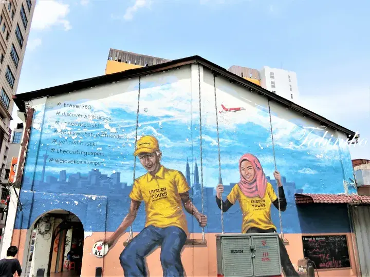 Mural of two people in yellow Unseen Tours shirts on a swing against a blue sky backdrop with Kuala Lumpur skyline.