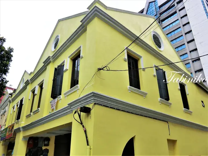 Bright yellow heritage shophouse with black shutters and grey trim, standing next to a modern high-rise building.