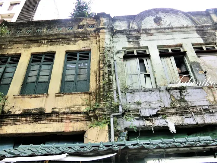 Weathered facade of a traditional shophouse with green louvered windows and peeling paint, showing signs of age and decay.