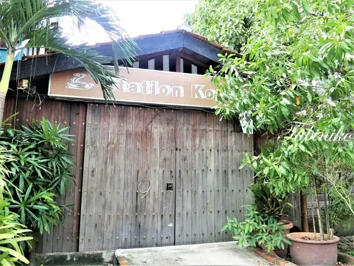 コーヒーカップのロゴが入った看板と、緑豊かな植物に囲まれた「Station Kopi」の木製ゲート。