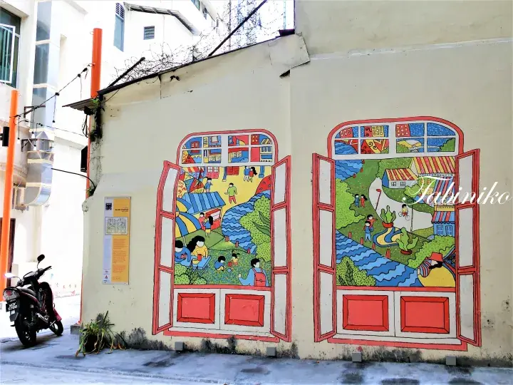 Colorful mural depicting a riverside scene painted on a cream wall in a Malaysian street, featuring a motorcycle parked nearby.