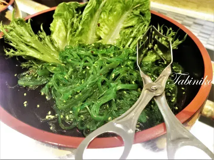 A wooden bowl filled with green seaweed salad and lettuce, accompanied by metal tongs.
