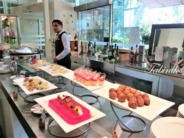 Buffet spread featuring a pink dessert, fried snacks, and fruit platter at a hotel with a waiter in the background.