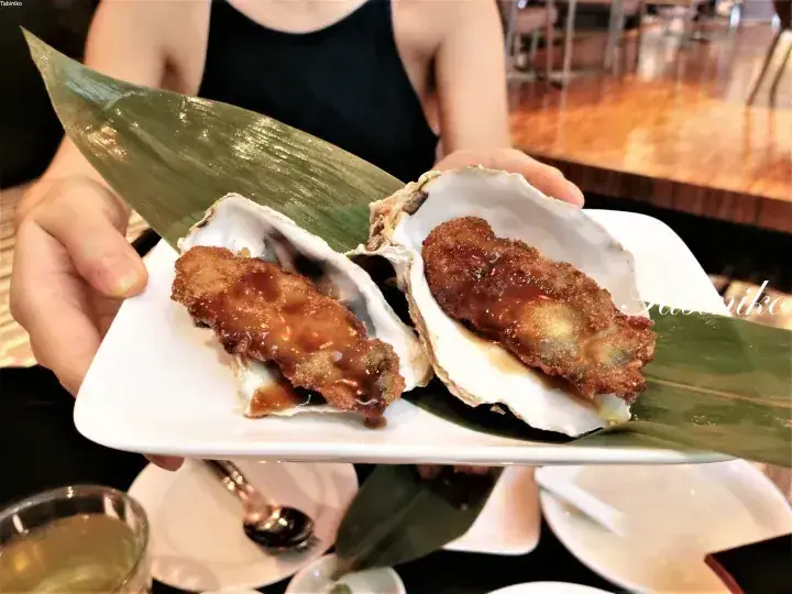 A person holds a plate with two fried oysters on the half shell, garnished with a green leaf.