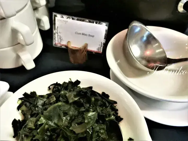 A bowl of dark leafy greens labeled Clam Mee Soup on a buffet table with serving utensils.
