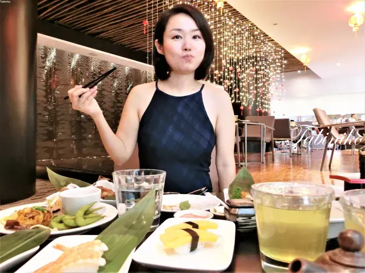 寿司と緑茶を前に食事をする女性、背景には装飾的な吊り下げライトが輝くレストランのシーン。