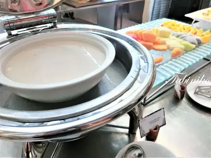 Creamy soup in a white bowl on a stainless steel chafing dish at a buffet, with fresh fruit platter in background