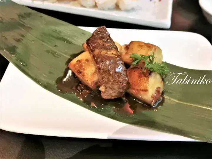 Grilled meat and potatoes served on a banana leaf with sauce and garnish, featuring the Tabiniko watermark.