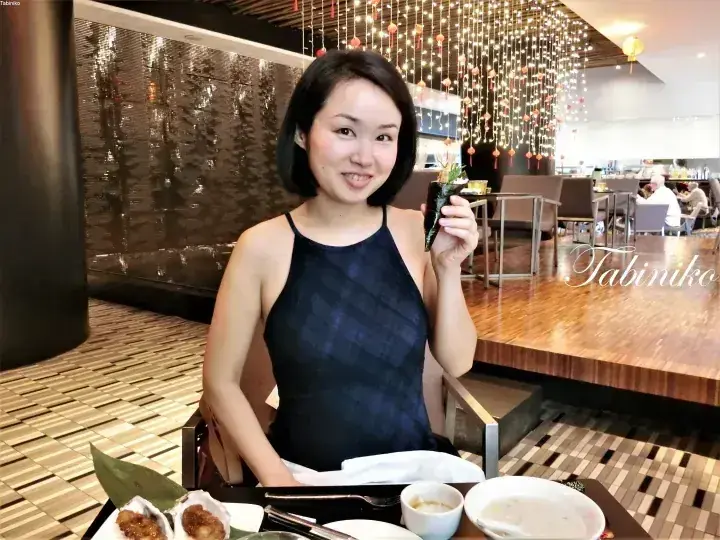 A woman smiles while holding a piece of sushi at a restaurant with a decorative water wall and hanging lights.