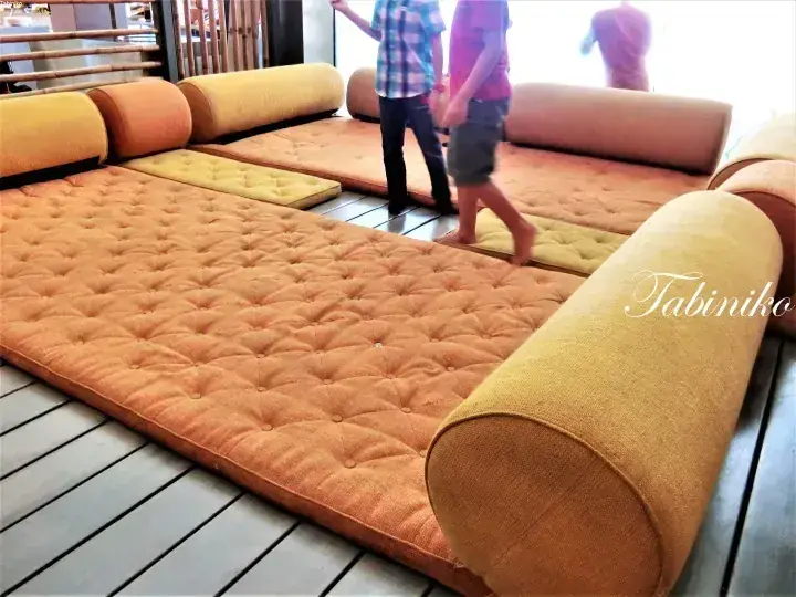 Large orange tufted floor cushions and cylindrical pillows arranged on a wooden deck at a resort.