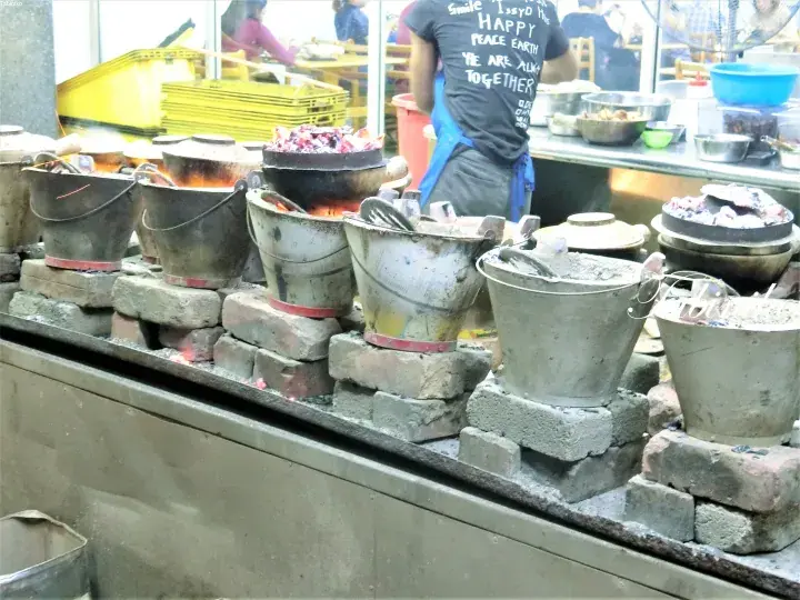Traditional clay pots simmering over brick stoves at a bustling Malaysian street food stall.