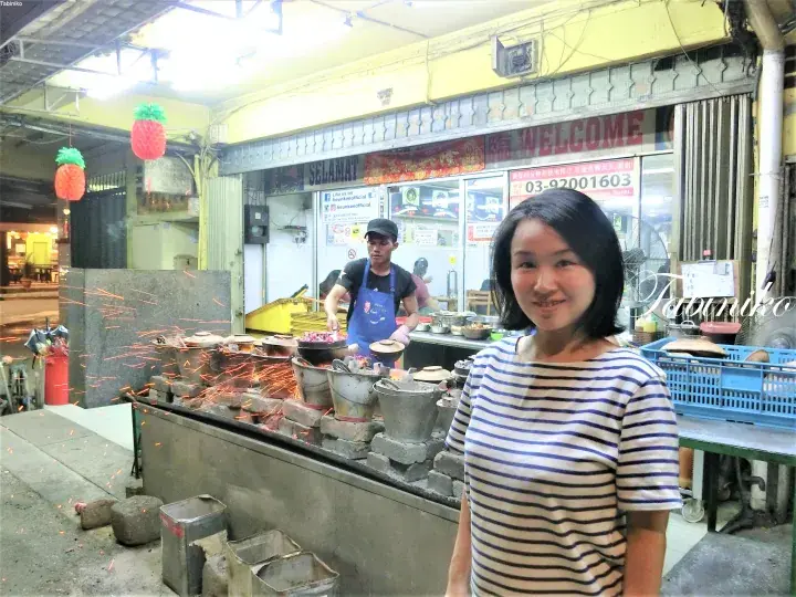 A woman stands smiling in front of a street food stall with sparks flying from a wok at night.
