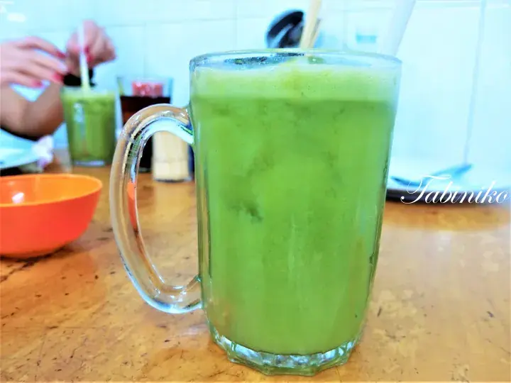 A tall glass mug filled with a vibrant green smoothie on a wooden table, with a person drinking a similar beverage in the background.